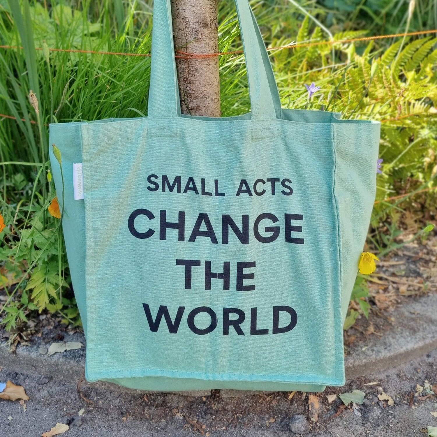 sustainable grocery bag multi pocket with 6 sleeves inside made of organic green cotton with the sentence: small acts change the world hanging on a wood stick